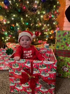 a baby is sitting in a christmas gift box with presents under the tree and smiling