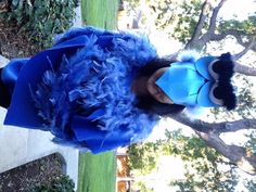 a woman wearing a blue feathered dress standing in front of a tree with her hands on her hips