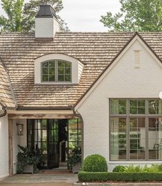 a white house with two large windows and a brown shingled roof, surrounded by greenery