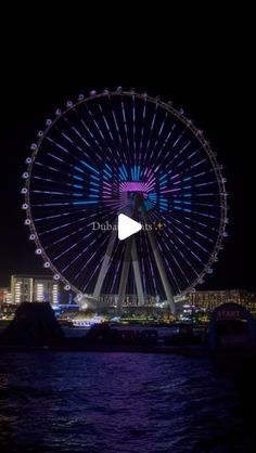 the ferris wheel is lit up at night in front of some buildings and water with lights on