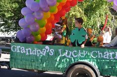 children in a parade float with balloons and shamrocks on the back, while adults watch