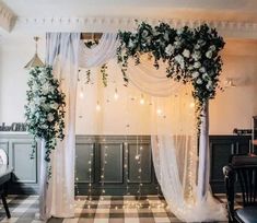 an indoor wedding ceremony with white flowers and greenery on the back wall, draped in sheer fabric