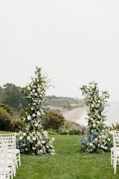 an outdoor ceremony set up with white chairs and flowers on the grass by the ocean