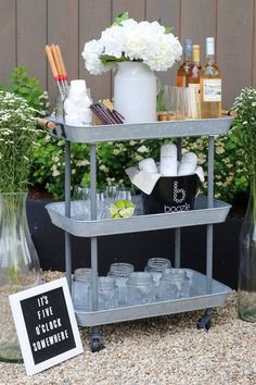 an outdoor bar cart with bottles and glasses on it, next to some white flowers