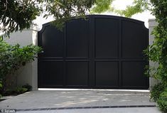 a large black gate in front of a white brick building with trees and bushes around it