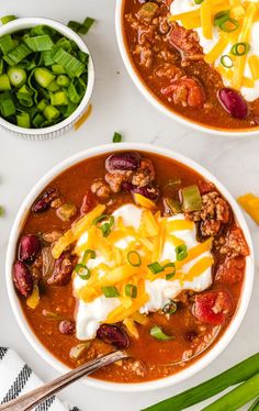 two bowls of chili with sour cream and cheese on top, surrounded by green onions