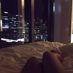 a woman laying in bed looking out the window at cityscape and skyscrapers