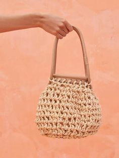 a hand holding a woven straw bag against a pink wall with the handle extended to it's side