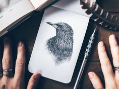 a person's hand is holding a pen and drawing a bird on a notebook