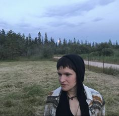 a woman standing in the middle of a field with trees and grass behind her, wearing a hoodie