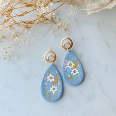 a pair of blue earrings with flowers painted on them and gold earwires next to dried plant