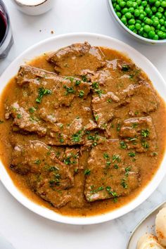 a white plate topped with meat covered in gravy next to bowls of peas