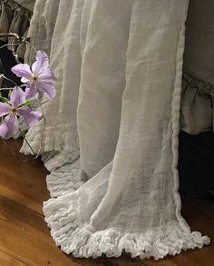 a bed with white ruffled bedspread and purple flowers in the corner next to it