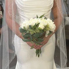 a woman in a wedding dress holding a bouquet of white flowers and greenery on her left hand