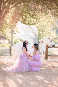 two women in dresses sitting next to a white horse with angel wings on its back