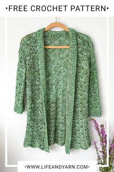 a green crochet jacket hanging on a wall next to a vase with flowers