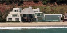 a large white house sitting on top of a sandy beach next to the ocean in front of a cliff