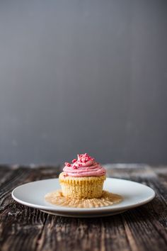 a cupcake with pink frosting on a white plate sitting on a wooden table