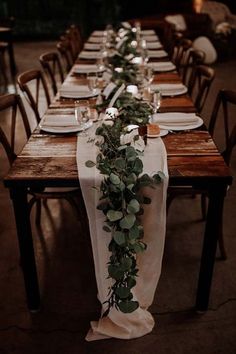 the long table is set with white plates and place settings, along with greenery