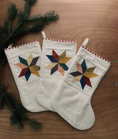 two white christmas stockings with colorful stars on them sitting next to a pine tree branch