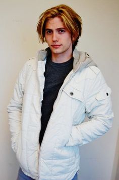 a young man is standing in front of a white wall wearing a jacket and jeans