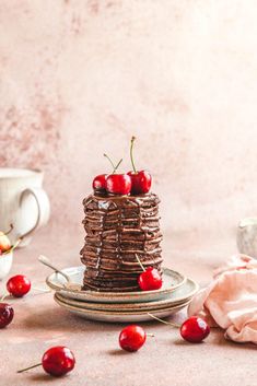 a stack of chocolate pancakes with cherries on top and coffee mugs in the background