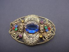 A delightful vintage Czech brooch - a large oval brooch set with a larger blue centre glass stone surrounded by n intricate filigree metal and claw set with a large blue glass centre stone surrounded by bright multi coloured hlass stones inan ornate metal work setting. The brooch is finished with an old  c clasp.  This is a nice large brooch that measures 2 & 1/2 inches (6.3 cm) across. The brooch is in good vintage condition with no damage, just a loose old c clasp. I will happily combine posta Blue Oval Brooch For Gifts, Blue Oval Brooch, Blue Oval Brooch For Formal Occasions, Ornate Blue Brooch For Formal Occasions, Ornate Oval Filigree Brooches, Ornate Blue Brooches For Wedding, Ornate Blue Wedding Brooches, Rainbow Colours, Metal Work