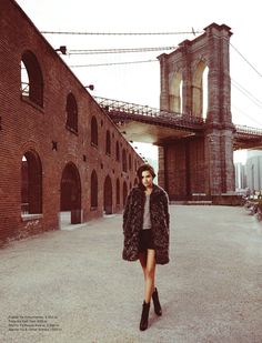 a woman is standing in front of an old brick building with a bridge behind her