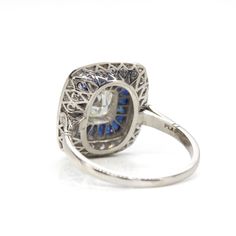 The Taylor ring is a piece handmade in platinum which centers one cushion old mine cut diamond of K color and VS1 clarity that weighs 0.75ctw. The center stone is surrounded by 20 natural calibrated cut sapphires that weigh 1.60ctw and 34 old mine cut diamonds that weigh 0.45ctw. Sapphire Platinum Diamond Ring Cushion Cut, Sapphire Cushion Cut Diamond Ring In Platinum, Platinum Sapphire Cushion Cut Diamond Ring, Sapphire Cushion Cut Diamond Ring, Sapphire Color Cushion Cut Diamond Ring, Fine Jewelry Platinum Cushion Cut Sapphire Ring, Cushion Cut Sapphire Ring With Diamond Accents, Cushion Cut Sapphire Diamond Ring, Diamond Cut Sapphire Ring With Cushion Shape