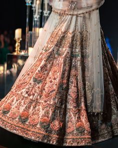 A true masterpiece that is an infusion of majestic details and glides beautifully into a floor-sweeping lehenga. The set has a lehenga, a blouse, a dupatta and a veil. A stunner lehenga tailored decked on a soft beige hue, in opulent raw silk with delicate 3D embroideries with organza flowers, pearls, salli, cutdana, and studs tassels. The sleeveless blouse showcases a contemporary deep sweetheart neckline with handcrafted detailing and a back hook closure. Paired with a heavy net dupatta done with cut-work detailing and intricate embroidery. The lehenga features an attached cancan and the blouse has padding. Finished with a veil, this mesmerizing silhouette defines extreme opulence. Dry-clean only if required. Slight variation in color is possible due to digital photography. Hand Embellished Lehenga For Navratri With Traditional Drape, Traditional Draped Hand Embellished Lehenga For Navratri, Hand Embellished Lehenga For Navratri, Floor-length Sherwani With Sheer Dupatta For Reception, Designer Raw Silk Lehenga With Intricate Embroidery, Semi-stitched Hand Embellished Raw Silk Lehenga, Hand Embellished Anarkali Lehenga For Festivals, Anarkali Hand Embellished Lehenga For Festivals, Anarkali Style Hand Embellished Lehenga For Diwali