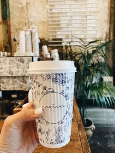 a hand holding a coffee cup on top of a wooden table next to a plant