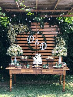 a table topped with cakes and flowers under a wooden wall covered in greenery,