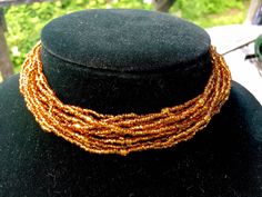 "Bead Choker with 12 strands of golden glass beads Small seed beads with a larger barrel bead about every 3\" Larger faceted bead fits in loop to close Adjustable size 13.5\" - 15.5\" 1.25\" wide Condition - very good" Bohemian Gold Choker With Colorful Beads, Handmade Multi-strand Gold Beaded Bracelets, Handmade Gold Multi-strand Beaded Bracelets, Adjustable Double Strand Gold Beads, Bohemian Gold Beaded Choker, Gold Double Strand Beaded Necklaces With Tiny Beads, Gold Double Strand Beaded Necklace With Tiny Beads, Gold Choker With Colorful Round Beads, Brown Multi-strand Beaded Necklace With Gold Beads