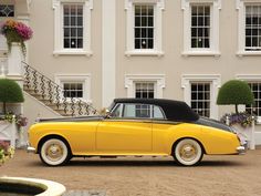 an old fashioned yellow car parked in front of a large white building with windows and balconies