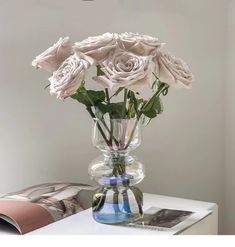 some white roses in a clear vase on a small table with magazines and a lamp