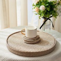 a white coffee cup sitting on top of a round place mat next to a vase with flowers