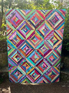 a colorful quilt hanging from the side of a tree in front of some bushes and trees