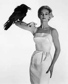 a woman in a white dress holding a black bird on her arm and looking at the camera