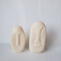 two white candles sitting next to each other on a white tablecloth covered surface, one is shaped like a face and the other has a nose