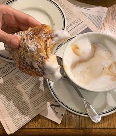 a person holding a pastry in their hand over a cup of coffee and newspaper on the table