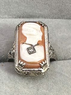 For your consideration, a 14K Art Deco, white gold cameo ring. This beautiful ring is hallmarked as 14K and guaranteed to be such. There is no makers mark. The cameo is a carved shell that is prong set. The setting measures 20.8 mm x 11.6 mm. The cameo's subject is wearing a traditional 1920s wavy hairstyle and is wearing a white gold chain with a 2-point diamond pendant.The setting is very typical of art deco styling with a floral motif all the way around the top of the ring  that continues around the sides of the ring. This fabulous ring is a size 7 1/2 weighs 3.27 g. It is a fabulous piece of history with a lot more history to be made! Antique Diamond Cabochon Rings, Fine Carved Diamond Jewelry, Carved Diamond Fine Jewelry, Art Deco White Gold Jewelry With Cabochon, Fine Jewelry Diamond Intaglio, Fine Diamond Intaglio Jewelry, Fine Jewelry Diamond With Intaglio Detail, Art Deco Cabochon Jewelry For Anniversary, Antique Diamond Intaglio Jewelry