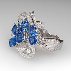 This beautiful cluster style ring is accented with one (1), prong set, oval mixed cut natural sapphire, two (2), prong set, triangular mixed cut natural sapphire and three (3), prong set, pear mixed cut natural sapphires. The sapphires are bordered with four (4), prong set, round brilliant cut diamond and thirty-five (35), bead set, round brilliant/single cut diamonds, bordered with milgrain edging. Engraved details accent the shoulders of the shank. The ring measures 20.3mm at the top, rises 10.4mm above the finger, tapering to 3.8mm wide and 1.3mm thick at the base of the shank. The ring is currently a size 6.75. Luxury Multi-stone Cluster Sapphire Ring, Luxury Multi-stone Sapphire Cluster Ring, Elegant Multi-stone Sapphire Cluster Ring, Elegant Sapphire Multi-stone Cluster Ring, Fine Jewelry Sapphire Cluster Ring With Rose Cut Diamonds, Platinum Multi-stone Sapphire Wedding Ring, Fine Jewelry Pear-shaped Gemstone Cluster Ring, Fine Jewelry Sapphire Ring Marquise Cut, Cluster Sapphire Ring In Fine Jewelry Style