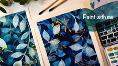 two paintings with blue and yellow leaves on them next to paintbrushes, watercolors and paper