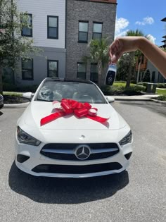 a white car with a red bow on the hood and someone holding a cell phone in front of it