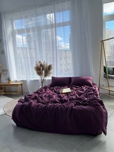 a large bed with purple sheets and pillows in front of two windows, next to a potted plant