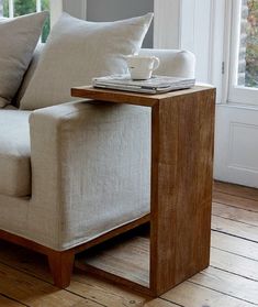 a couch with pillows and a coffee cup on it next to a wooden table in front of a window