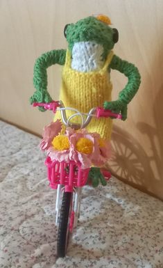 a knitted frog riding a pink bicycle with flowers on the front wheel and handlebars