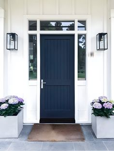 two planters with purple flowers are in front of a blue door on a white house