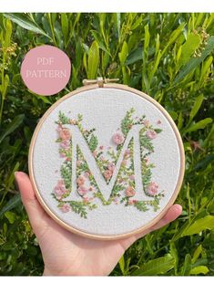 a hand holding up a cross stitched monogrammed letter m with pink flowers