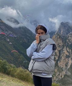 a woman standing on top of a mountain next to a lush green hillside covered in flying kites